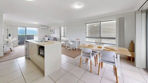 A modern kitchen with a central island, a dining area with a wooden table and white chairs, and a large window with blinds. - Cute & Cosy with Great Location (Moreton Bay City)