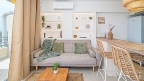 Un salon moderne comprenant un canapé, une table basse et un coin repas. On y trouve des étagères ornées d’objets décoratifs et un luminaire suspendu.