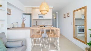 Une cuisine compacte avec un coin repas, des meubles blancs, un luminaire suspendu en rotin et un grand miroir au mur.