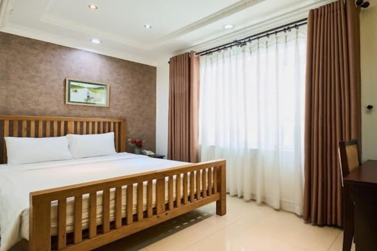 A hotel room with a wooden bed, a nightstand, a desk, and a window with curtains.