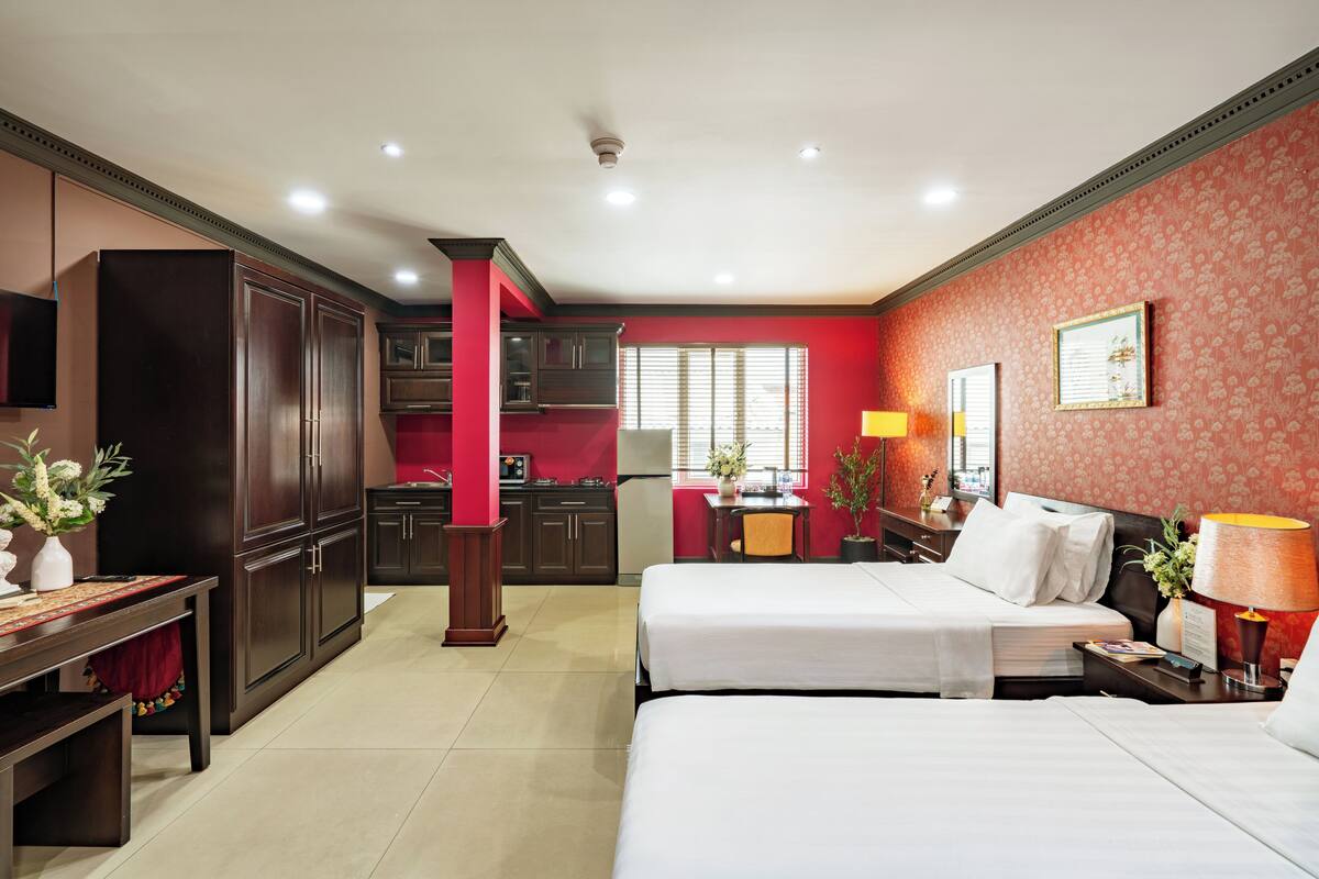 Ein Hotelzimmer mit zwei Betten, einem Schreibtisch, einem Stuhl, einer kleinen Küchenzeile und einer roten Akzentwand.