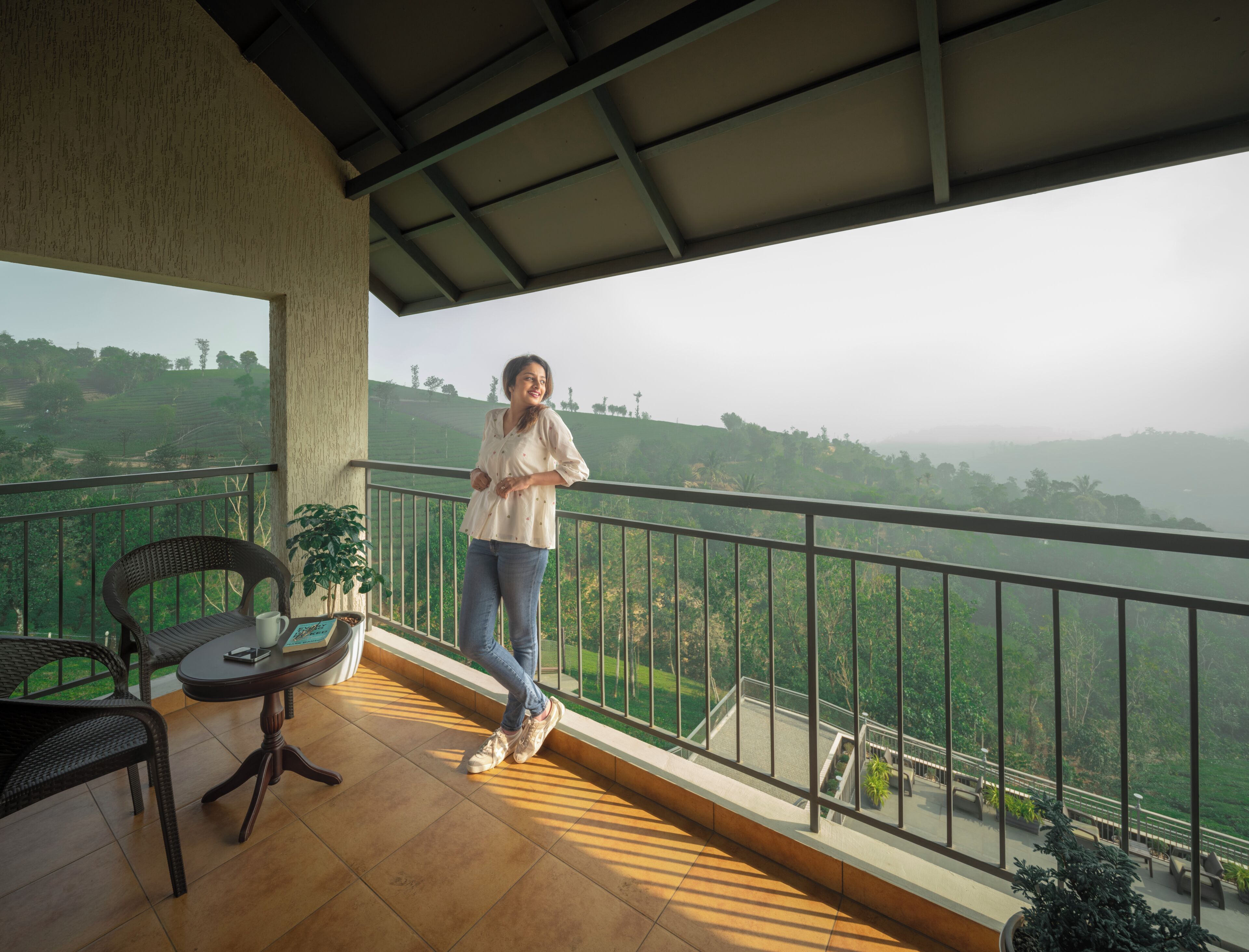 A balcony with a view of a hilly landscape, a wicker chair, a small table with a book and a cup, and a person standing.