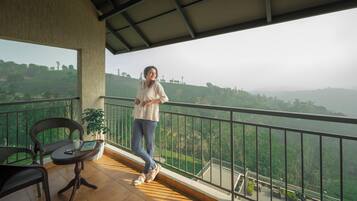A balcony with a view of a hilly landscape, a wicker chair, a small table with a book and a cup, and a person standing.