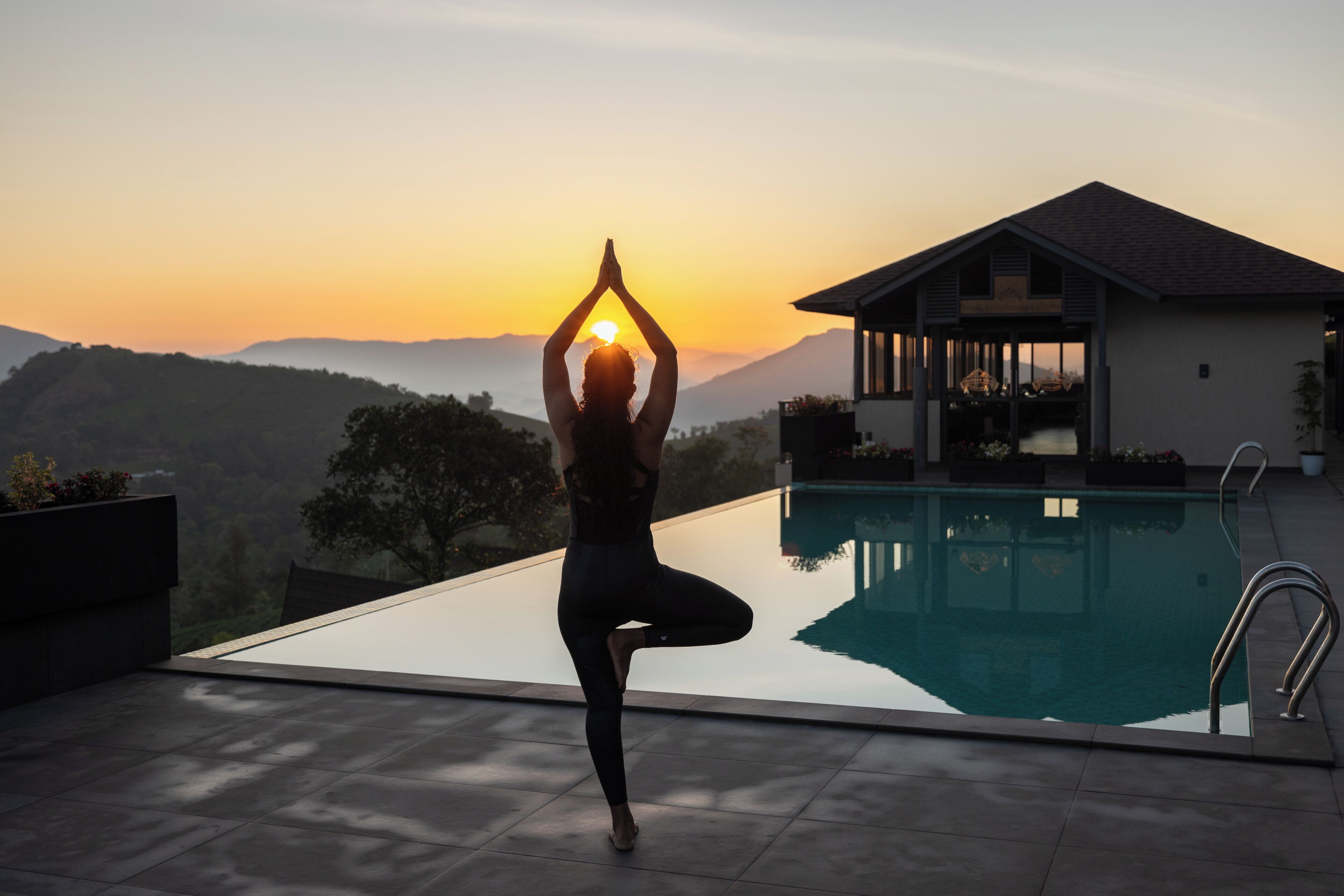 Una persona practicando yoga en una azotea con una piscina y una casa al fondo.