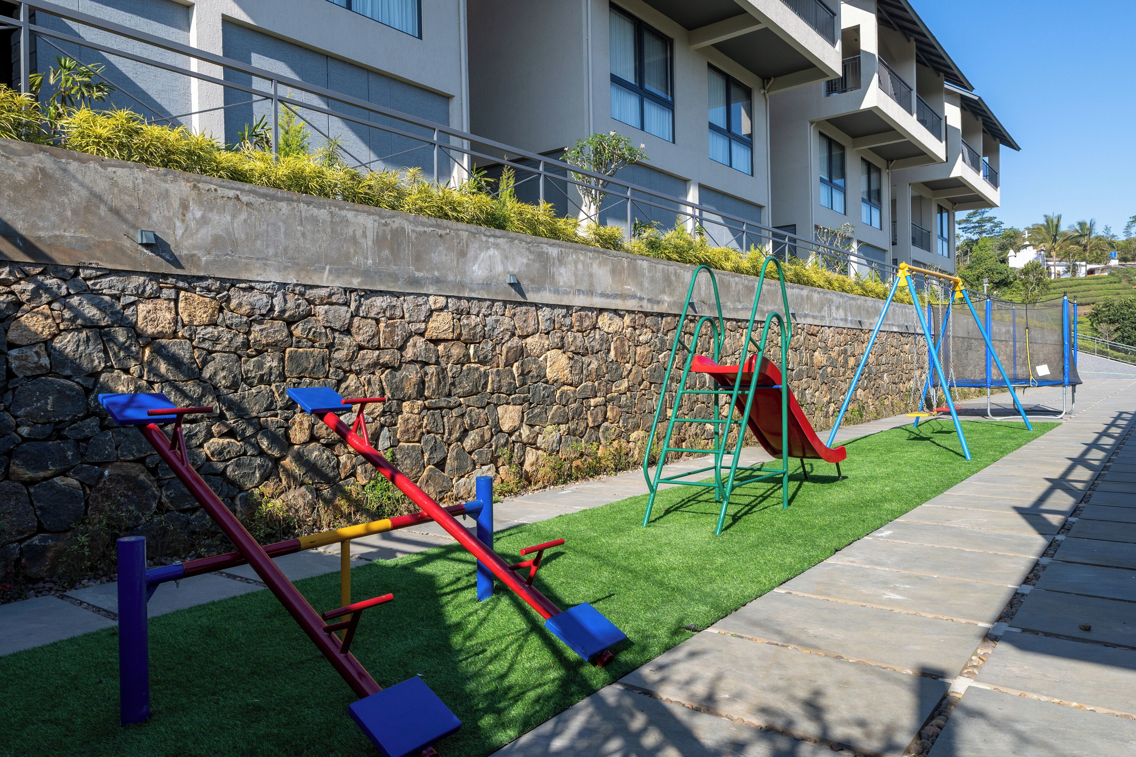 Columpios y juegos infantiles al aire libre, un muro de piedra y un edificio con balcones.