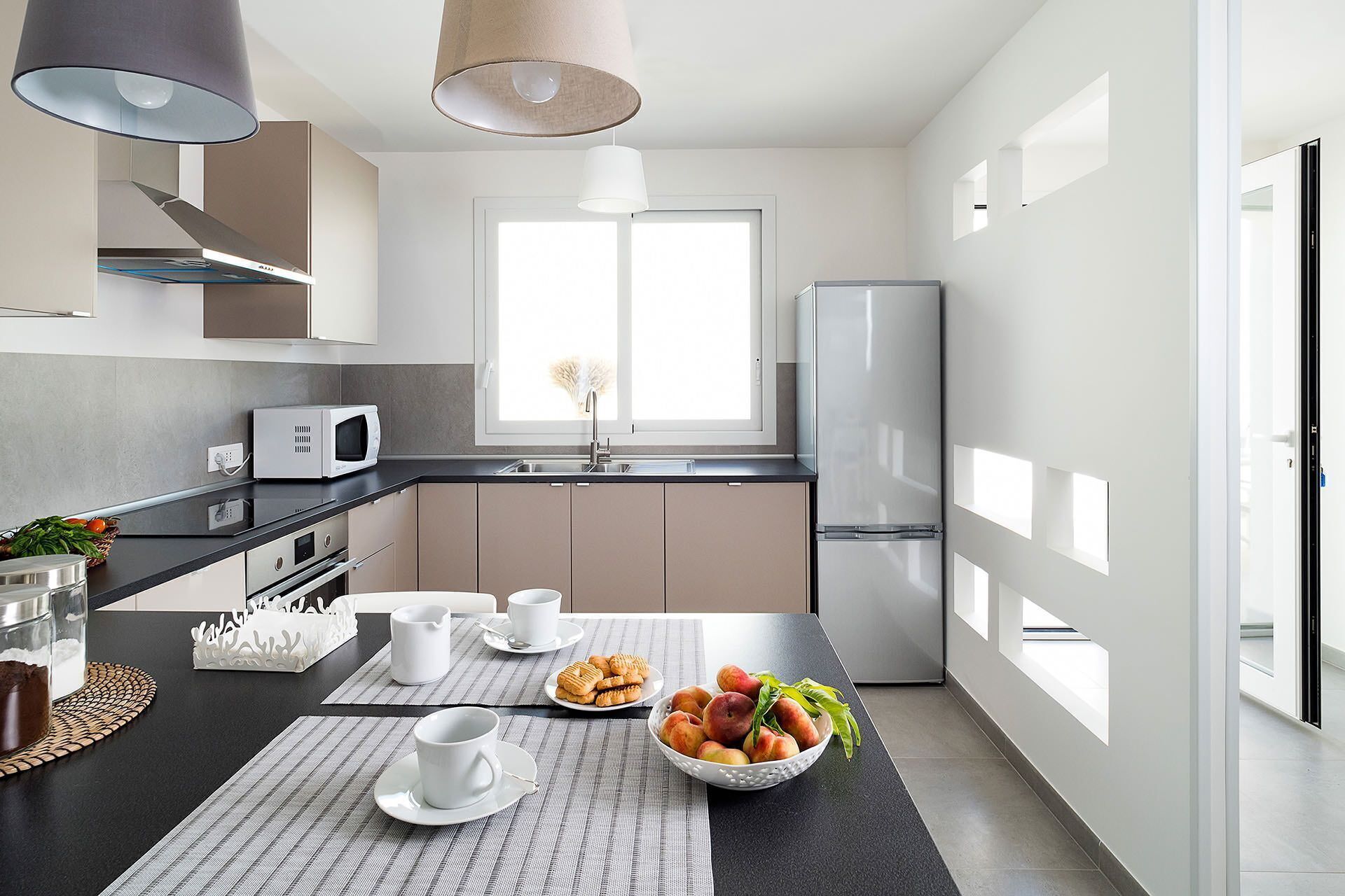 Een moderne keuken met een zwart aanrecht, witte kastjes, een magnetron, een spoelbak en een koelkast. Er zijn twee hanglampen, een raam met natuurlijk licht, en een tafel met kopjes, een fruitschaal en een mand met koekjes.