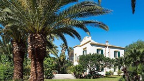 A white building with green shutters, surrounded by palm trees and a well-maintained garden. - Green Villa with heated pool - My Rental Homes (Salemi)