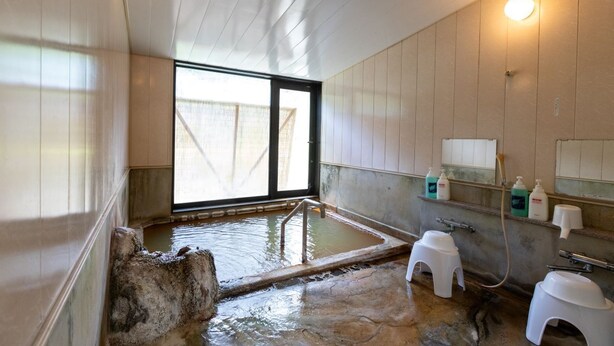 Un onsen japonais traditionnel, avec une source d’eau chaude naturelle entourée de pierres, des murs en bois et une fenêtre laissant entrer la lumière naturelle.