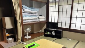 Une pièce avec un futon servant de lit, un téléviseur posé sur un meuble, un ventilateur et une étagère avec des couvertures pliées.