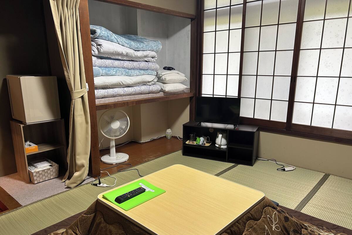 A room with a futon bed, a TV on a stand, a fan, and a shelf with folded blankets.
