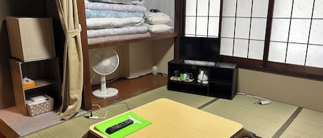 Une pièce avec un futon servant de lit, un téléviseur posé sur un meuble, un ventilateur et une étagère avec des couvertures pliées.