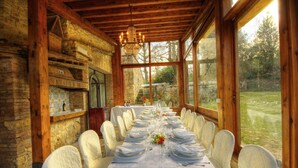 Una larga mesa de comedor dispuesta para una comida, con mantel blanco y sillas, en una habitación rústica de madera con ventanas amplias.