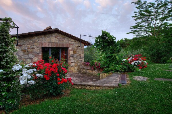 A stone cottage with a wooden door, surrounded by lush greenery and vibrant flowers. - My Rental Homes - Villa Incanto with pool offers an amazing view of Montepulicano (Montepulciano)