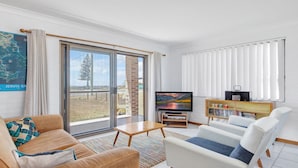 A modern living room with a large sliding glass door, a flat-screen TV, a bookshelf, and a comfortable seating area. - 2 Storey family home just steps away from the beach! (Culburra Beach)