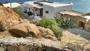 A coastal building with a stone pathway leading to a white structure, overlooking the sea. - My Rental Homes - Villa Zenas with stunning pool overlooking the Aegean sea. (Mykonos)