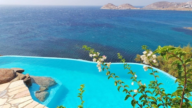 A pool with clear blue water, surrounded by rocks and plants, overlooking a calm sea and distant islands. - My Rental Homes - Villa Zenas with stunning pool overlooking the Aegean sea. (Mykonos)