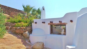 A white building with a small window, surrounded by greenery and a stone wall. - My Rental Homes - Villa Zenas with stunning pool overlooking the Aegean sea. (Mykonos)