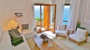 A living room with a sofa, armchair, and a coffee table. There is a view of the ocean through large windows. - My Rental Homes - Villa Zenas with stunning pool overlooking the Aegean sea. (Mykonos)