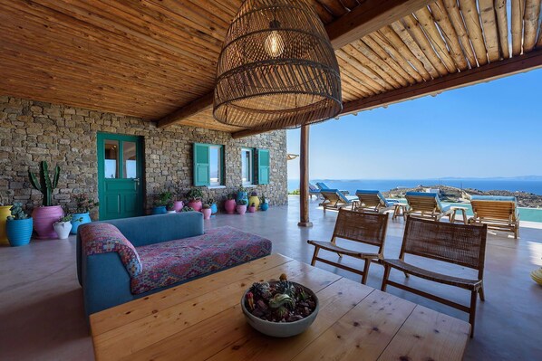 Quarto acolhedor com parede de pedra, teto de madeira e vista para o mar. Há plantas coloridas em vasos, um sofá azul e cadeiras de madeira.