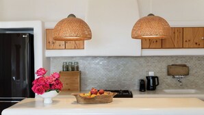A kitchen with two woven pendant lights, wooden cabinets, a white countertop, a vase of pink roses, and a fruit bowl. - My Rental Homes - Villa Joy with sea view, pool equipped with sunbeds and patio (Mykonos)