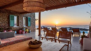 A cozy outdoor seating area with wicker furniture and a table, overlooking a sunset over the sea. - My Rental Homes - Villa Joy with sea view, pool equipped with sunbeds and patio (Mykonos)