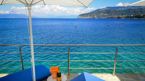 Un balcon avec vue sur la mer, une table avec une bouteille et des verres, et des chaises bleues avec des coussins.