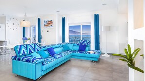 Un salon moderne doté d’un grand canapé d’angle, d’un coin repas avec des chaises transparentes et offrant une vue sur la mer.