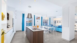Une cuisine moderne dotée d’un îlot central, d’un coin repas avec des chaises transparentes et d’une vue sur la mer.