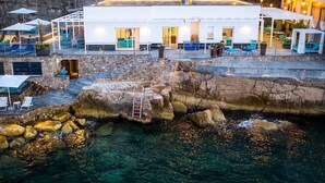 Un hôtel situé sur la côte, avec une façade blanche, des places assises en plein air et une vue sur le littoral rocheux.