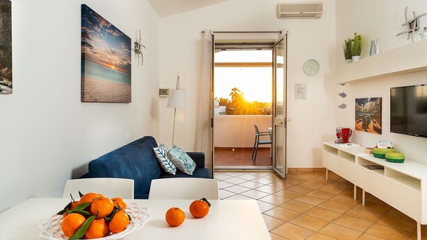 A modern living room with a sofa, a dining table with oranges, a TV, and a view of the sunset through an open door.