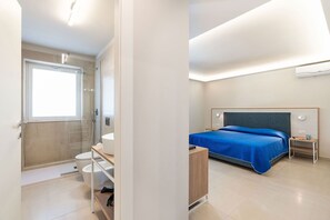 Ein modernes Hotelzimmer mit Dusche, einem Bett mit blauer Bettdecke, einem Nachttisch und einem Beistelltisch.