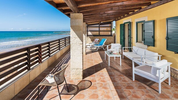 Un'area salotto esterna coperta, con mobili bianchi, un muro in pietra e vista sul mare.