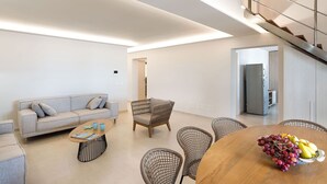 Un soggiorno moderno con divano, tavolino e poltrone in vimini. Si nota un tavolo da pranzo in legno con della frutta. Una scala conduce a un livello superiore.