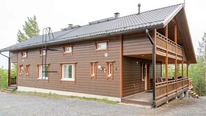 Una baita di legno a due piani con un balcone, un vialetto sterrato e uno sfondo boschivo.