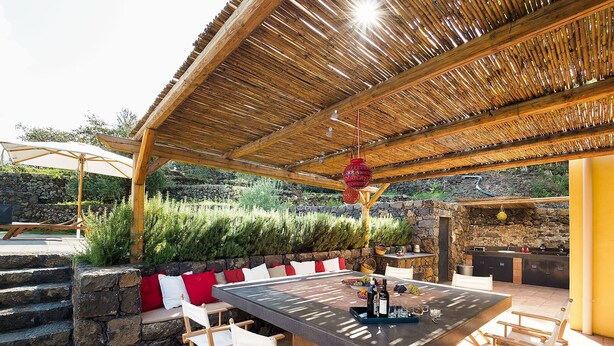 Un área de comedor al aire libre con pared de piedra, sillas de madera, mesa con cubierta de vidrio y techo de paja.
