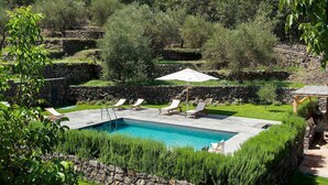 Piscina con sillones de descanso y una sombrilla, rodeada de muros de piedra y follaje.