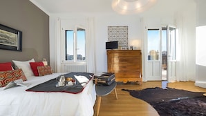 Une chambre d’hôtel avec un lit, une commode, un bureau sur lequel se trouve un plateau avec des articles pour le petit-déjeuner, et une vue sur la ville grâce à de grandes fenêtres.