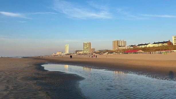 A beach with a calm water body reflecting the sky, buildings in the background, and a clear blue sky.