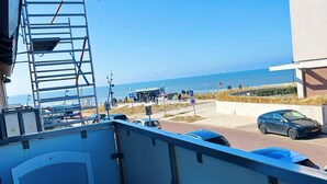 A balcony with a view of the beach, a parked car, and a building in the background.