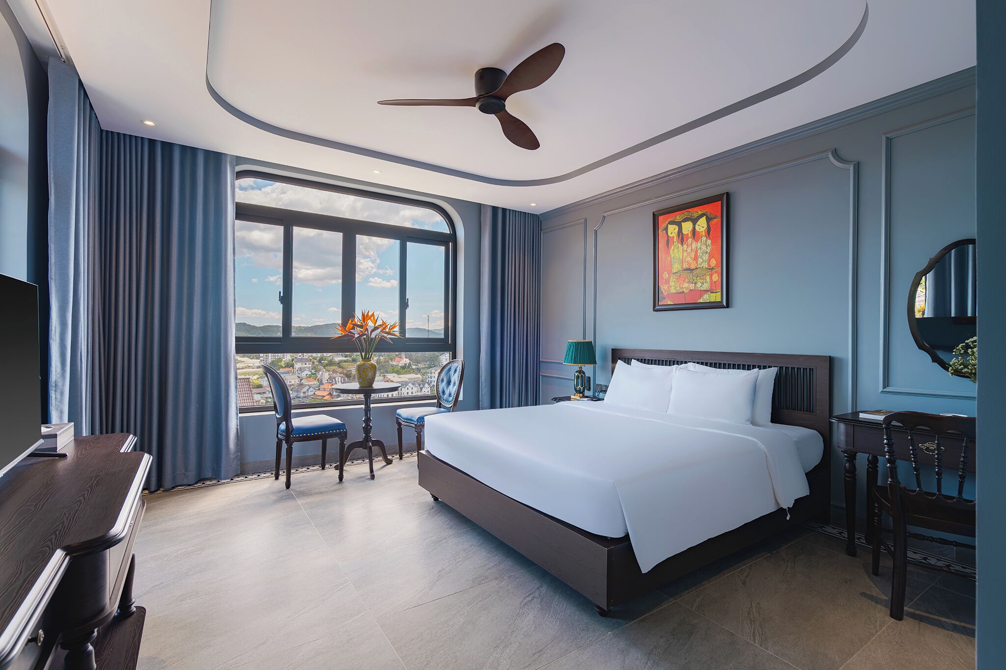 Quarto de hotel com uma cama grande, ventoinha no teto, vista para a cidade, mesa de jantar e uma cadeira.