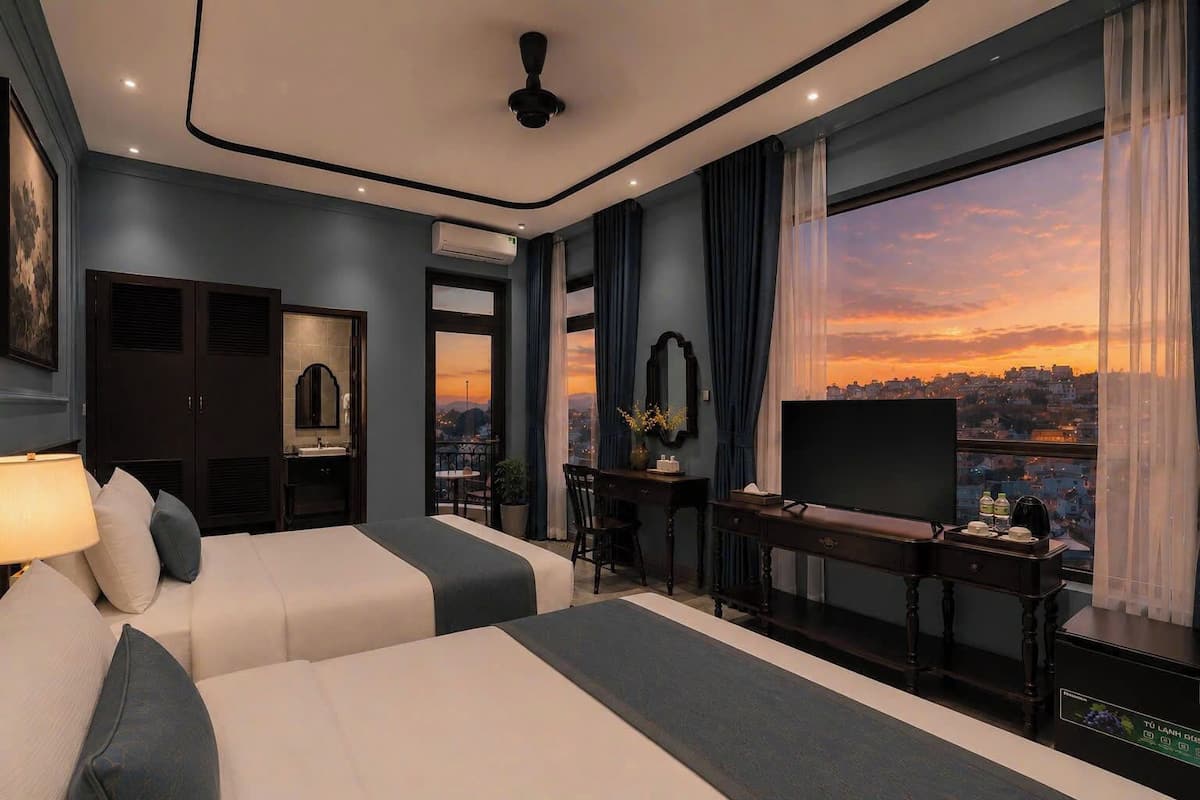 A hotel room with a large bed, a desk with a TV, a view of the city at sunset, and a balcony with a table and chairs.