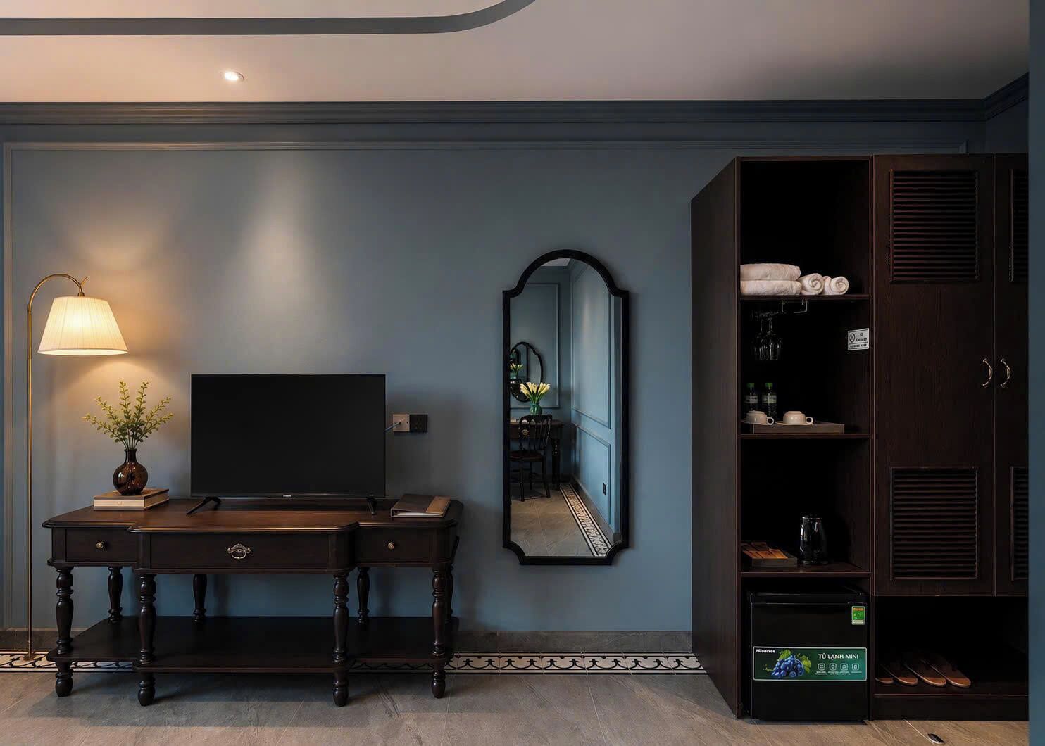 A hotel room with a dark wooden desk, a flat-screen TV, a mirror, and a wardrobe with shelves.