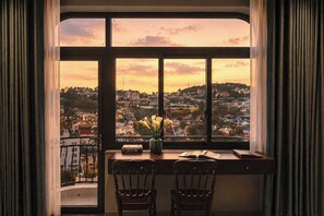 A room with a large window overlooking a cityscape at sunset, a desk with a vase of flowers, and two chairs. - Lumina Dalat Premium hotel (Da Lat)