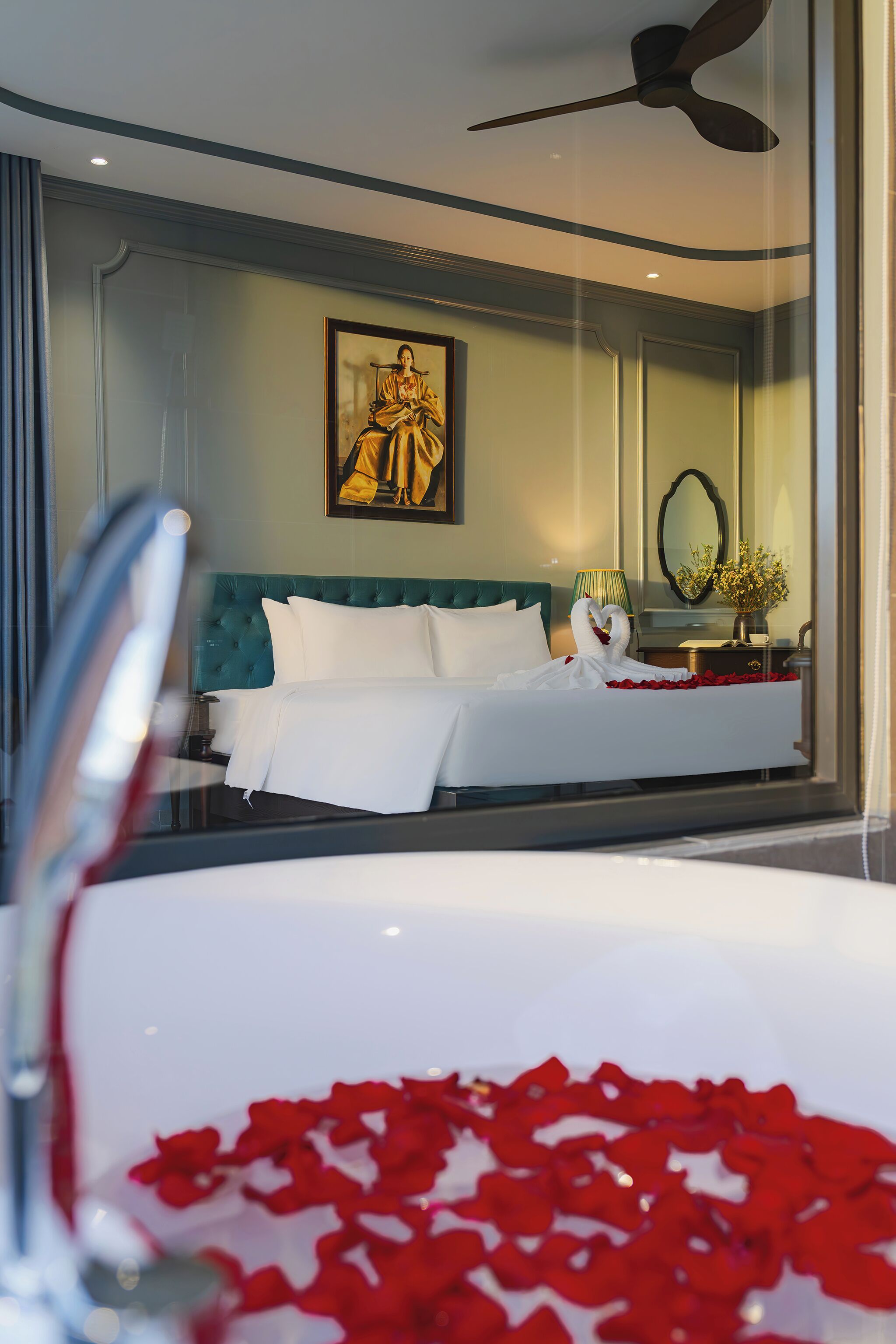 A hotel room with a bed, a ceiling fan, a framed artwork on the wall, and a vase with flowers on the nightstand.