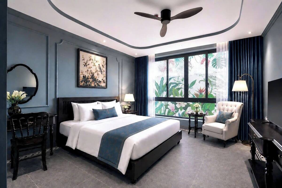 A modern hotel room with a large bed, a sitting area with a chair, a desk, a TV, and a large window with a view of greenery.