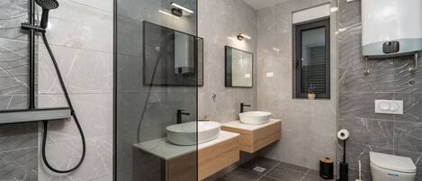 Un baño moderno con cabina de ducha de vidrio, dos lavamanos con armarios de madera, un inodoro y suelo de baldosas de mármol.
