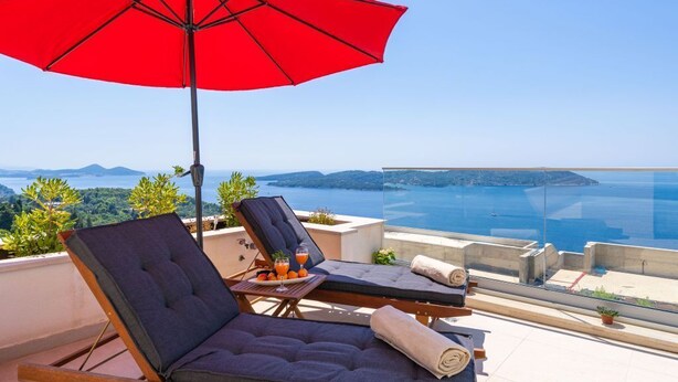 A sun lounger with a red umbrella, a table with drinks, and a scenic view of the sea and hills.