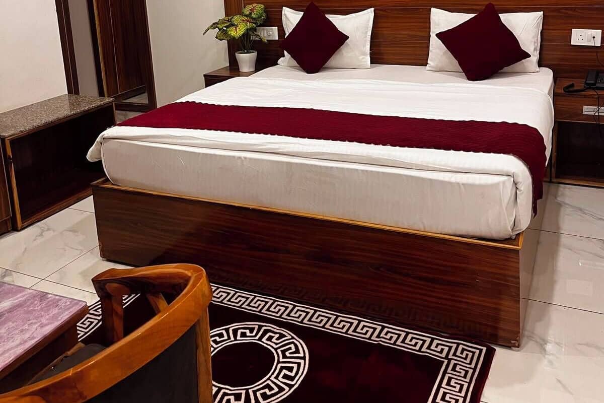 A neatly arranged bedroom with a wooden headboard, a bed with white and maroon bedding, a framed painting above the bed, a small table with a chair, and a patterned rug on the floor.