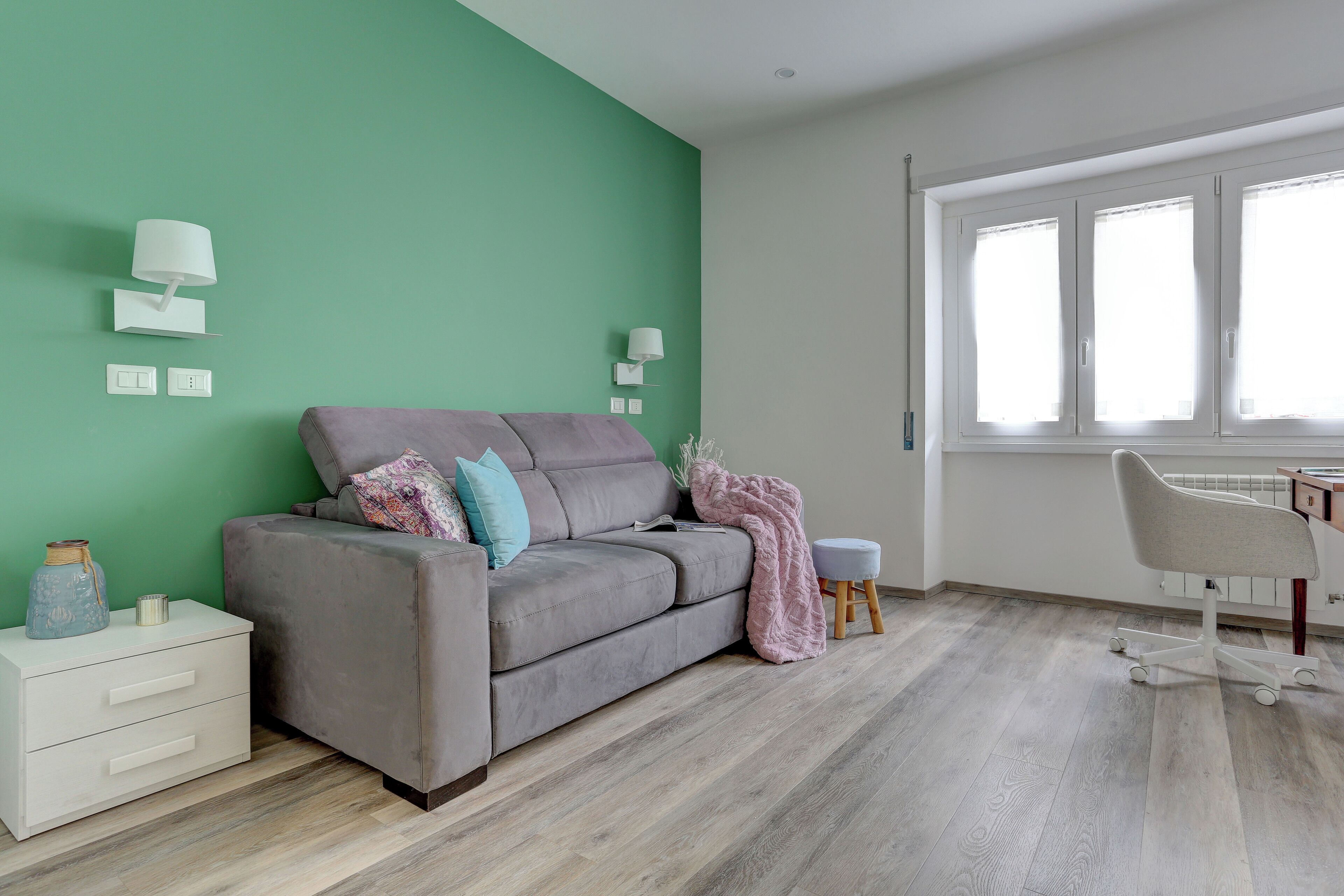 A modern living room with a grey sofa, a white nightstand, a small stool, and a desk with a chair.