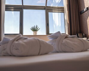 Una cama bien hecha con toallas, una planta en maceta en el alféizar y vista al cielo a través de la ventana.